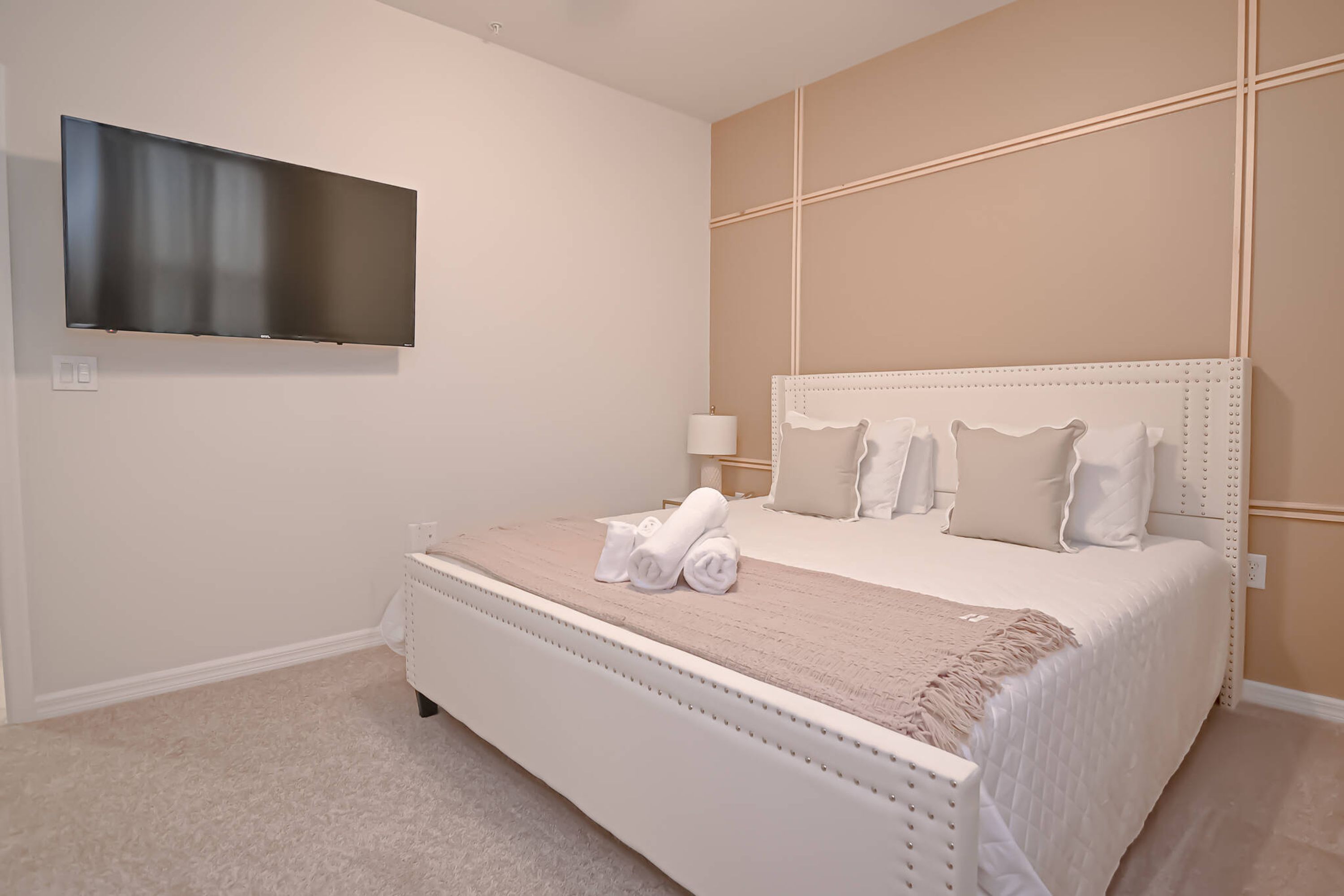 A bedroom with a large bed, a flat-screen TV mounted on the wall, and a bedside table with a lamp.