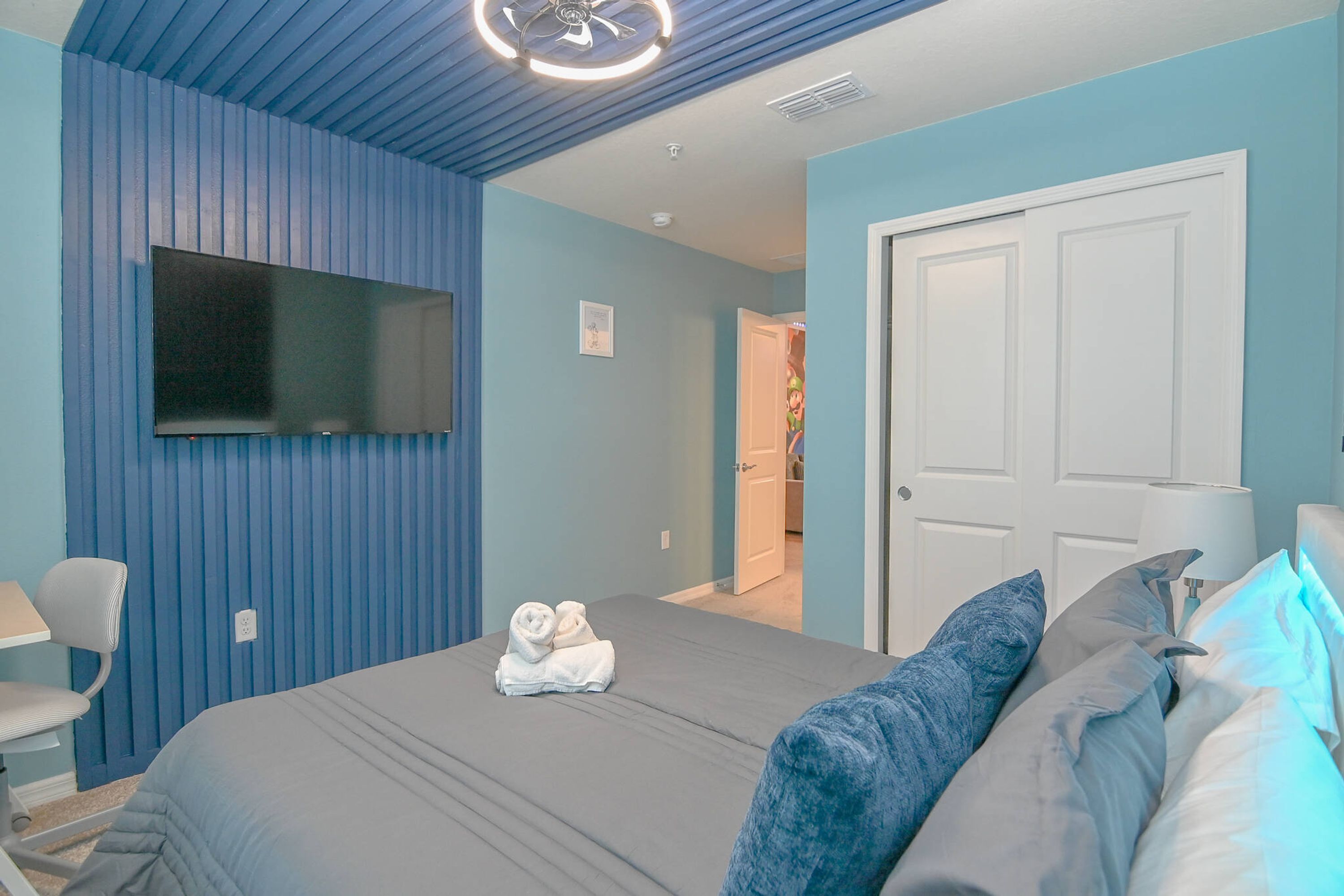 A bedroom with a bed, a TV mounted on the wall, a desk, and a chair.