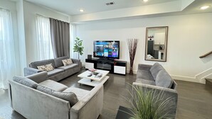 A modern living room with a sectional sofa, a flat-screen TV, a coffee table, and a large mirror on the wall. - ROOFTOP VIEWS OF NYC+FREE Parking+Washer&Dryer (Jersey City)
