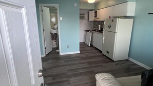 Eine moderne Küche mit weißen Schränken und einem Kühlschrank, ein Flur mit Lichtschalter sowie ein Wohnbereich mit einer Couch.