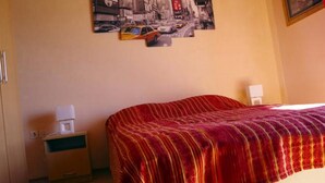 Une chambre d’hôtel avec un lit recouvert d’une couverture à rayures rouges et jaunes, une table de chevet avec une lampe et un tableau représentant un paysage urbain accroché au mur.