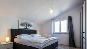 A modern bedroom with a black headboard, a bedside table with a lamp, a large window with curtains, and a painting on the wall.