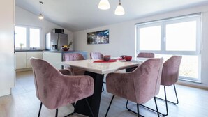 A modern kitchen with white cabinets, a dining table with pink chairs, pendant lights, and a large window.