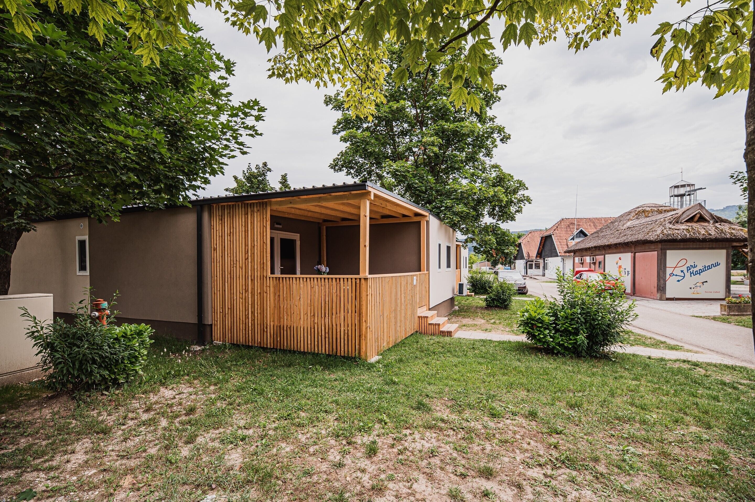A small, single-story wooden structure with a covered porch, surrounded by grass and trees.