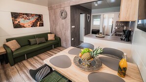 Ein modernes Wohnzimmer mit einem grünen Sofa, einem Esstisch mit Obst-Dekoration und einem durch eine offene Tür einsehbaren Küchenbereich.