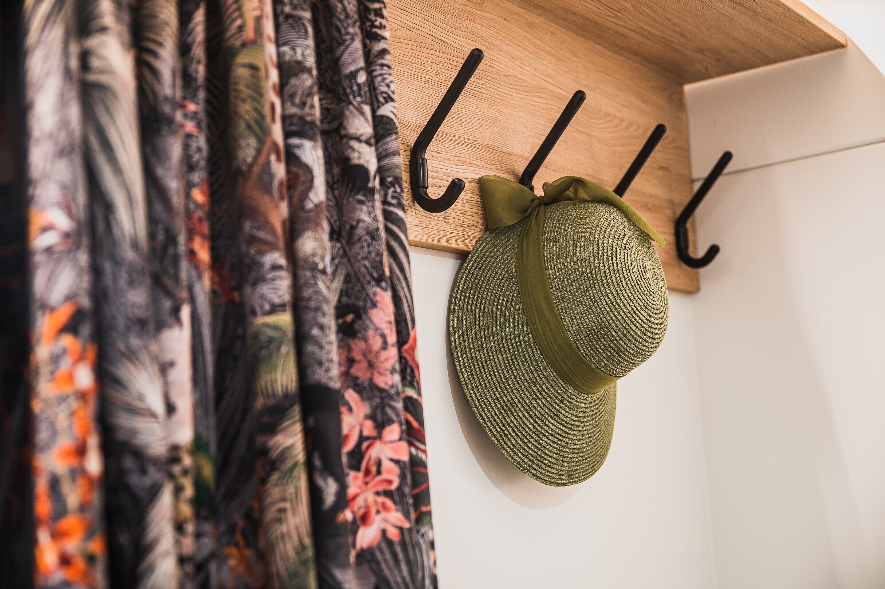 A hat hanging on a wooden hook with a floral curtain in the background.