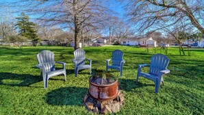 A grassy outdoor area with four Adirondack chairs arranged around a metal fire pit.