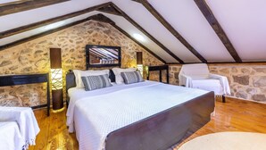 A bedroom with a stone wall, wooden beams, a bed with white bedding, a chair, and a small table.