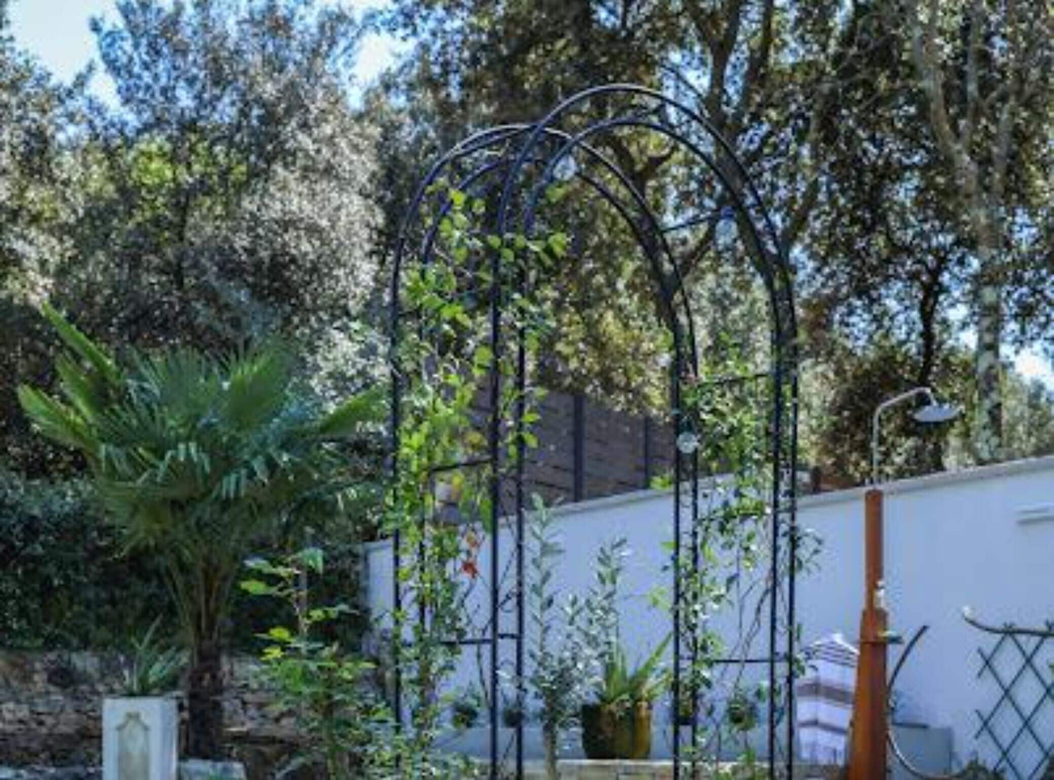 Un jardin agrémenté d’une arche en métal, de différentes plantes et, au fond, d’un bâtiment blanc.