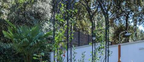 Un jardín con un arco de metal, diversas plantas y un edificio blanco al fondo.