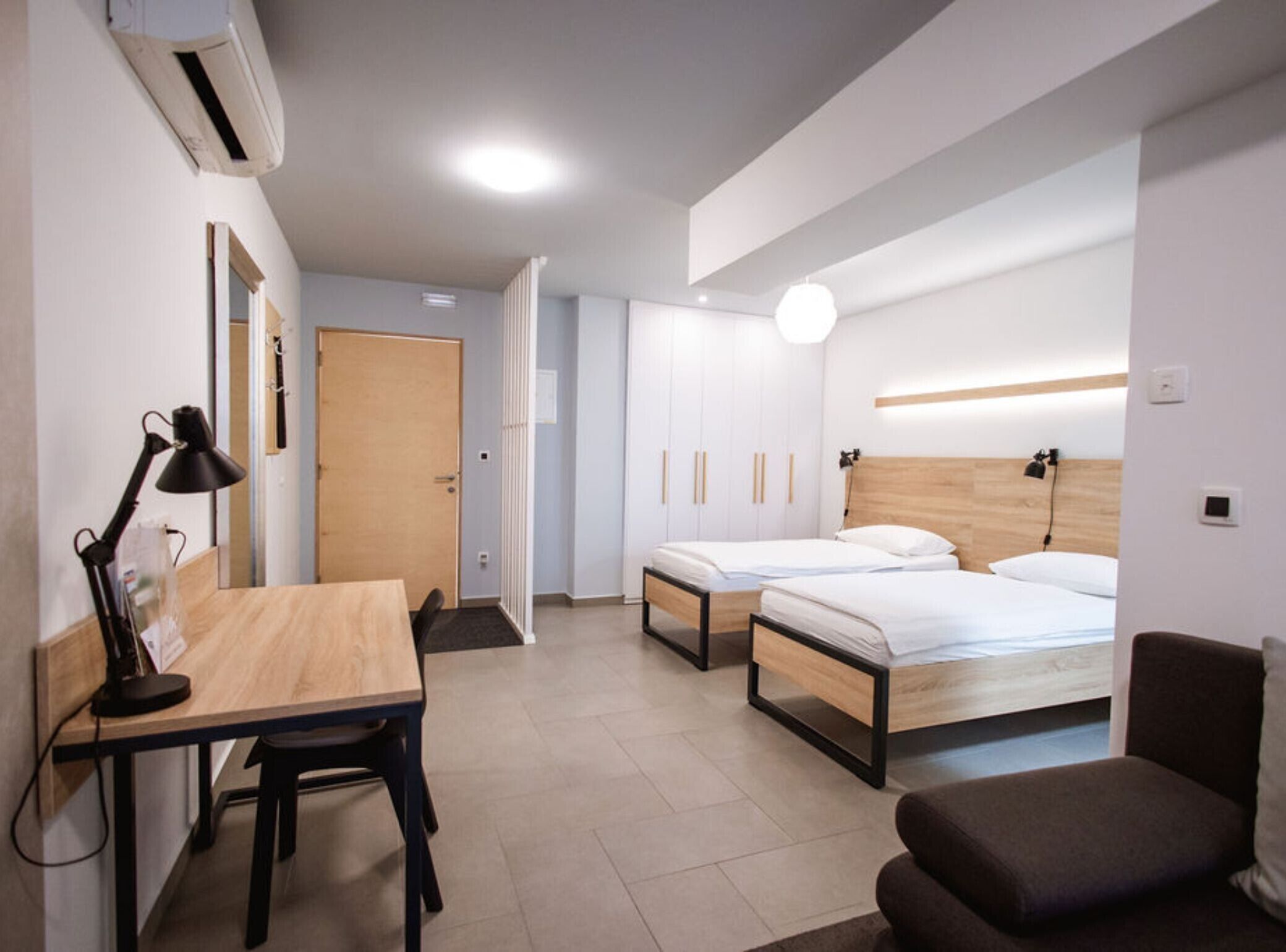 A modern hotel room with a wooden desk, two beds, a sofa, and a wardrobe.