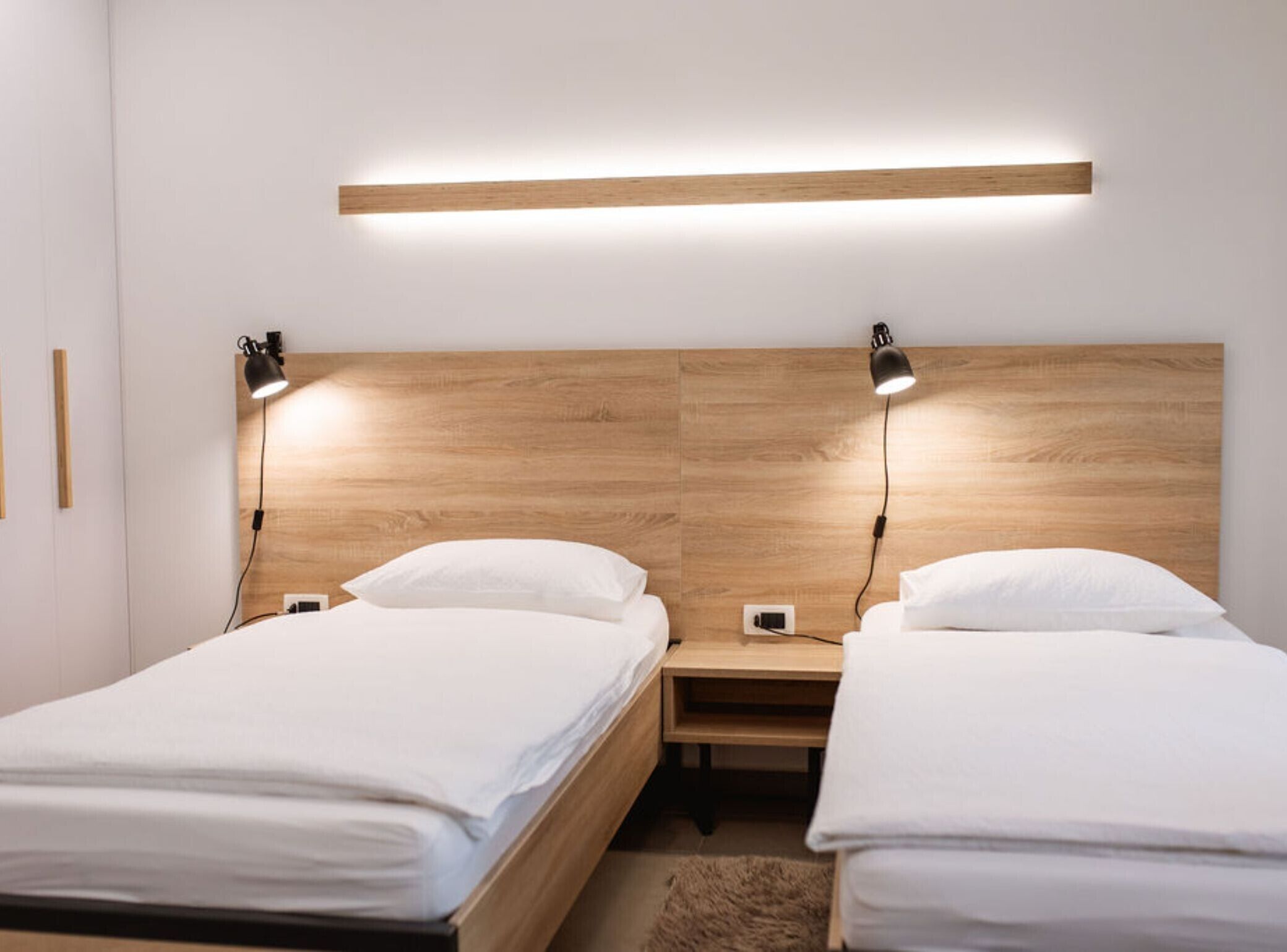 A modern bedroom with two single beds, wooden headboards, bedside lamps, and a built-in shelf above the beds.
