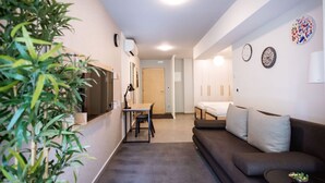 A compact living space with a sofa, a dining table, and a TV. There is a plant on the left and a door leading to another room. - Stylish Apartments with Osijek - Standard Studio (Osijek)