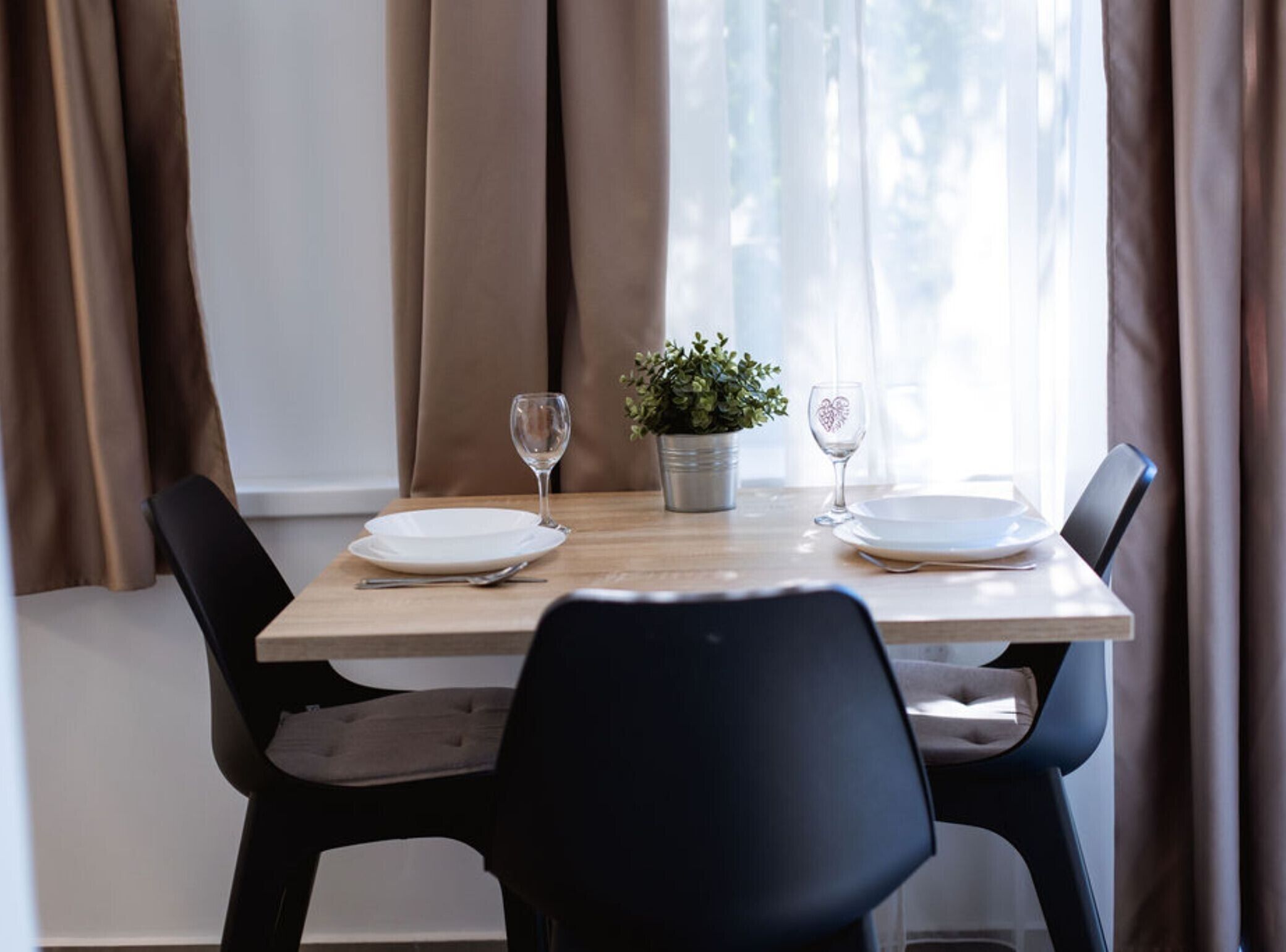 A dining table set with plates, cutlery, and wine glasses, with a potted plant and curtains in the background.