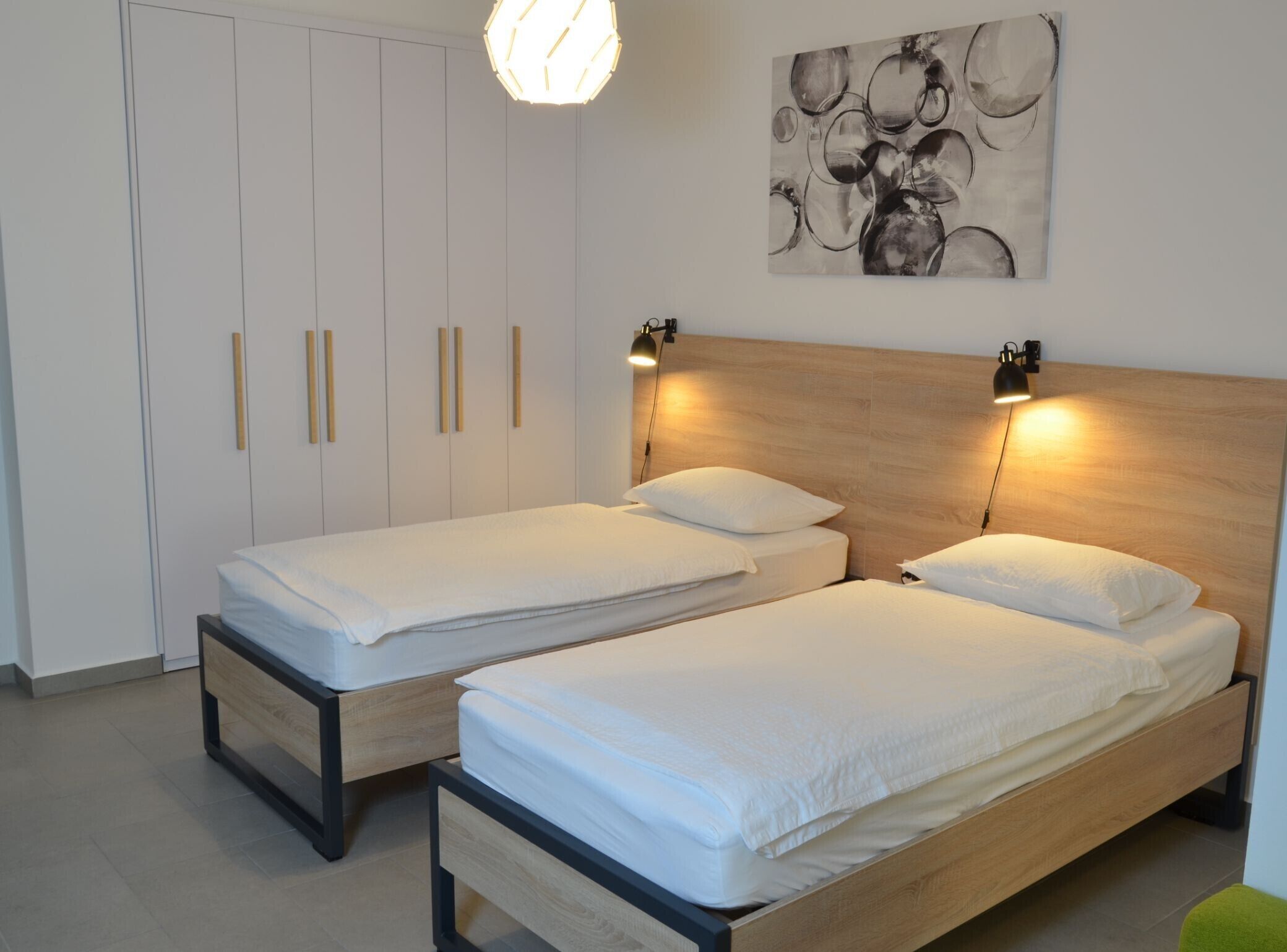 A bedroom with two single beds, a wooden headboard, a wall-mounted abstract painting, and a pendant light.
