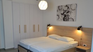 Een moderne slaapkamer met een houten bedframe, wit beddengoed, een hoofdbord met geïntegreerde leeslampjes, een wanddecoratie in abstractie en een kledingkast met verticale handgrepen.