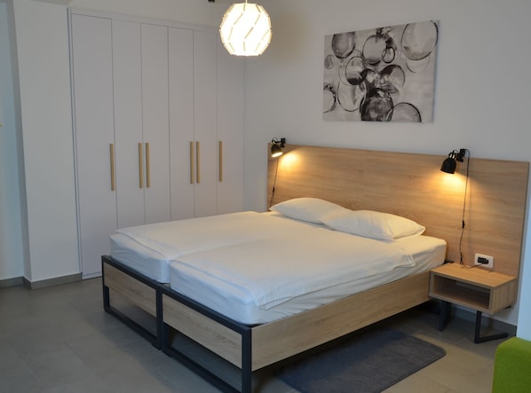 A modern bedroom with a wooden bed frame, white bedding, a headboard with integrated reading lights, a wall-mounted abstract art piece, and a wardrobe with vertical handles.