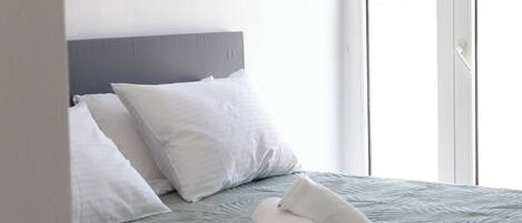 Un lit bien fait, avec une tête de lit grise, des oreillers blancs et une serviette blanche roulée posée sur une couverture grise à texture.