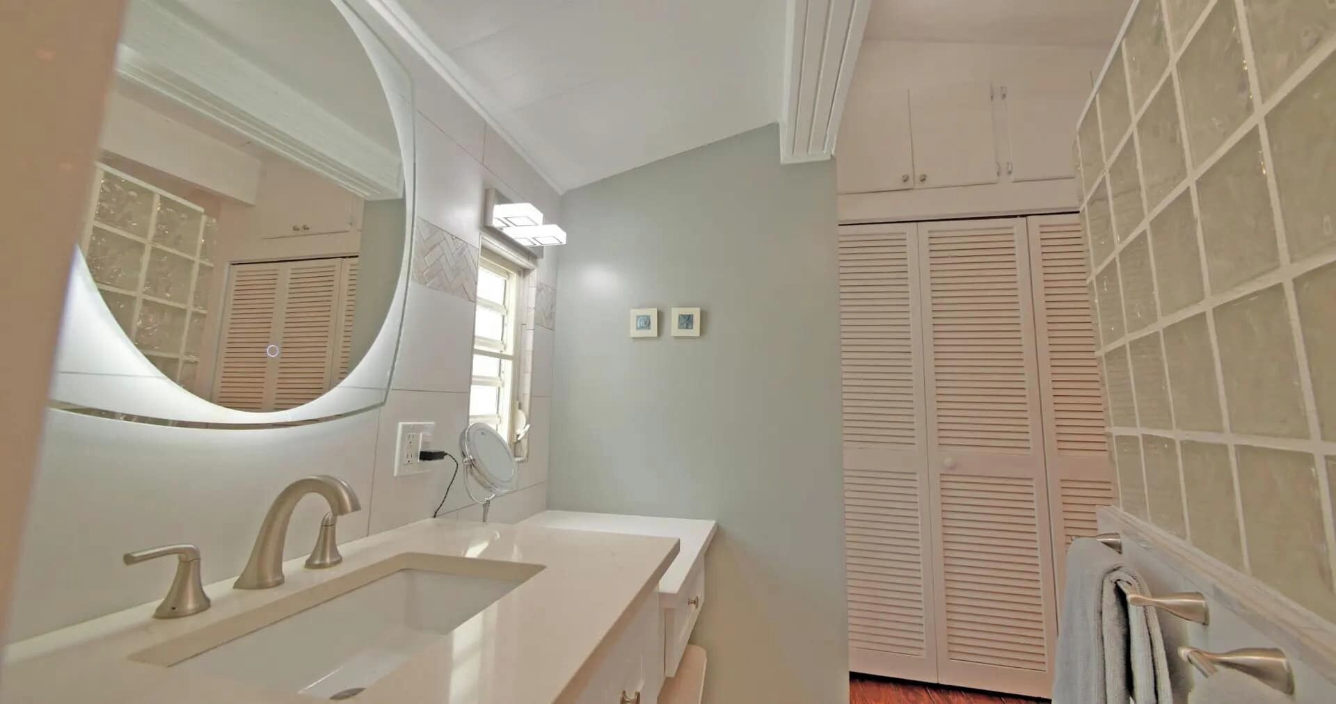 Un baño con un espejo grande, un lavabo y tocador. Hay dos cuadros enmarcados en la pared y una ventana con persianas.