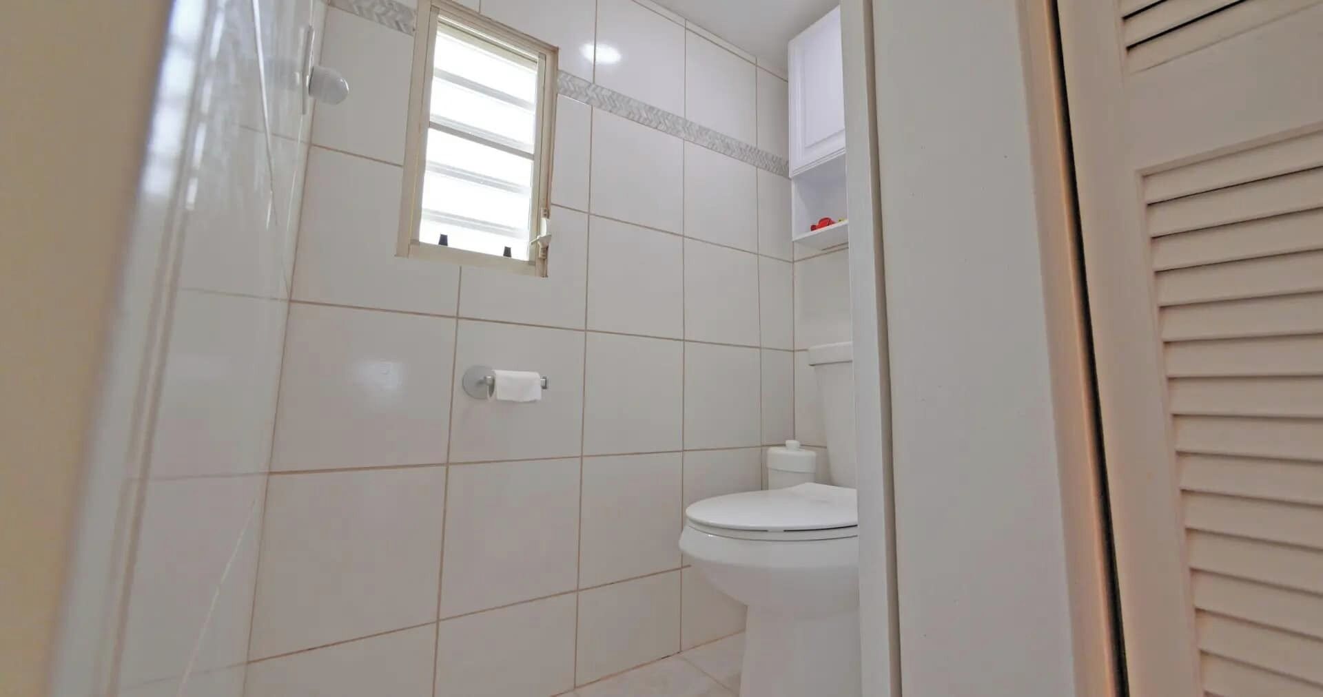 A bathroom with white tiles, a toilet, a window, and a towel dispenser.