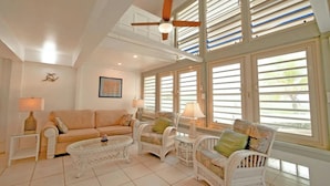 Amplia sala de estar con techo alto, ventanas grandes con contraventanas, ventilador de techo y un área de descanso confortable con un sofá y dos sillones de mimbre.