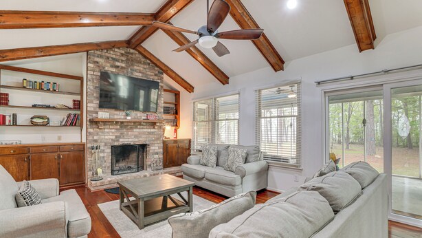A spacious living room with a fireplace, wooden beams, a ceiling fan, and large windows. - Patio + Pool Table: Lake Marion Getaway (Santee)