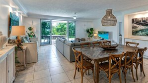 A spacious living area with a dining table and chairs, a sofa, and a large window offering a view of the outdoors. - Poinciana 012 Hamilton Island & Buggy (Whitsundays)