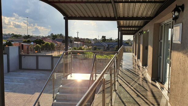 Un espacio exterior techado con barandilla de vidrio, un camino pavimentado y vistas del barrio circundante.