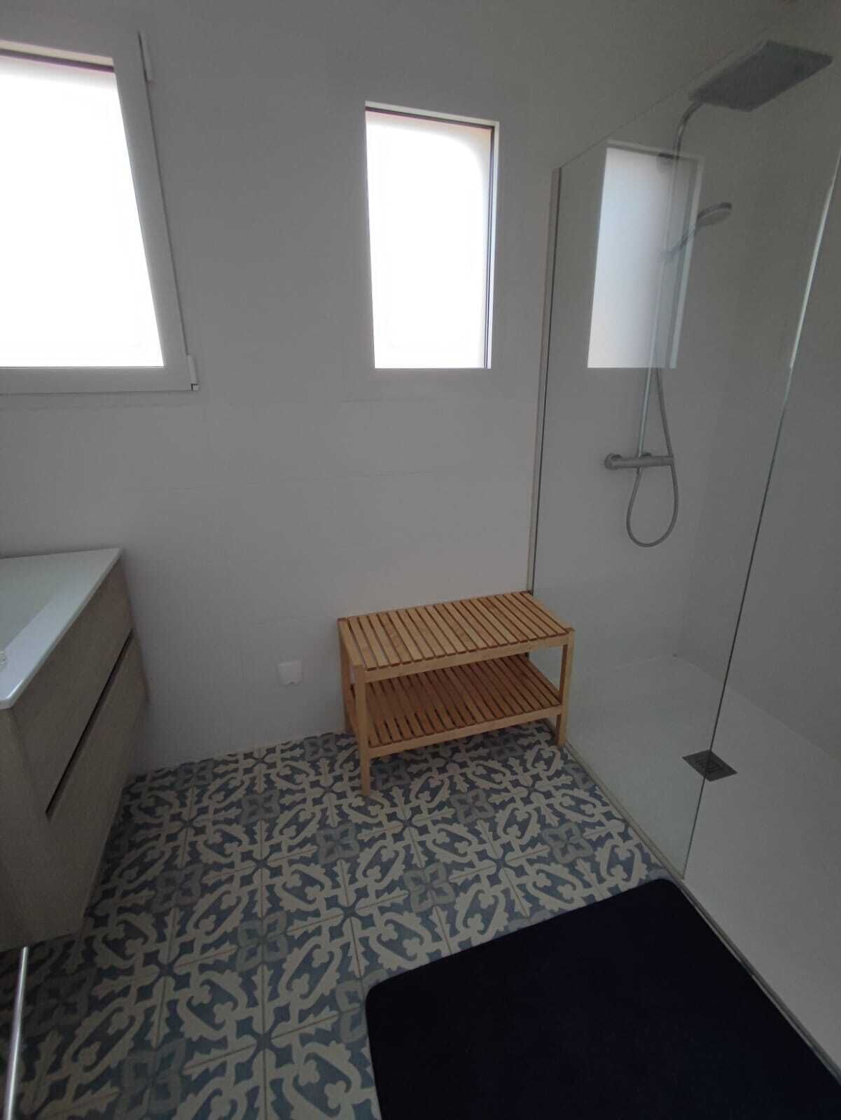 A small bathroom with a shower, two windows, a wooden shelf, and patterned floor tiles.