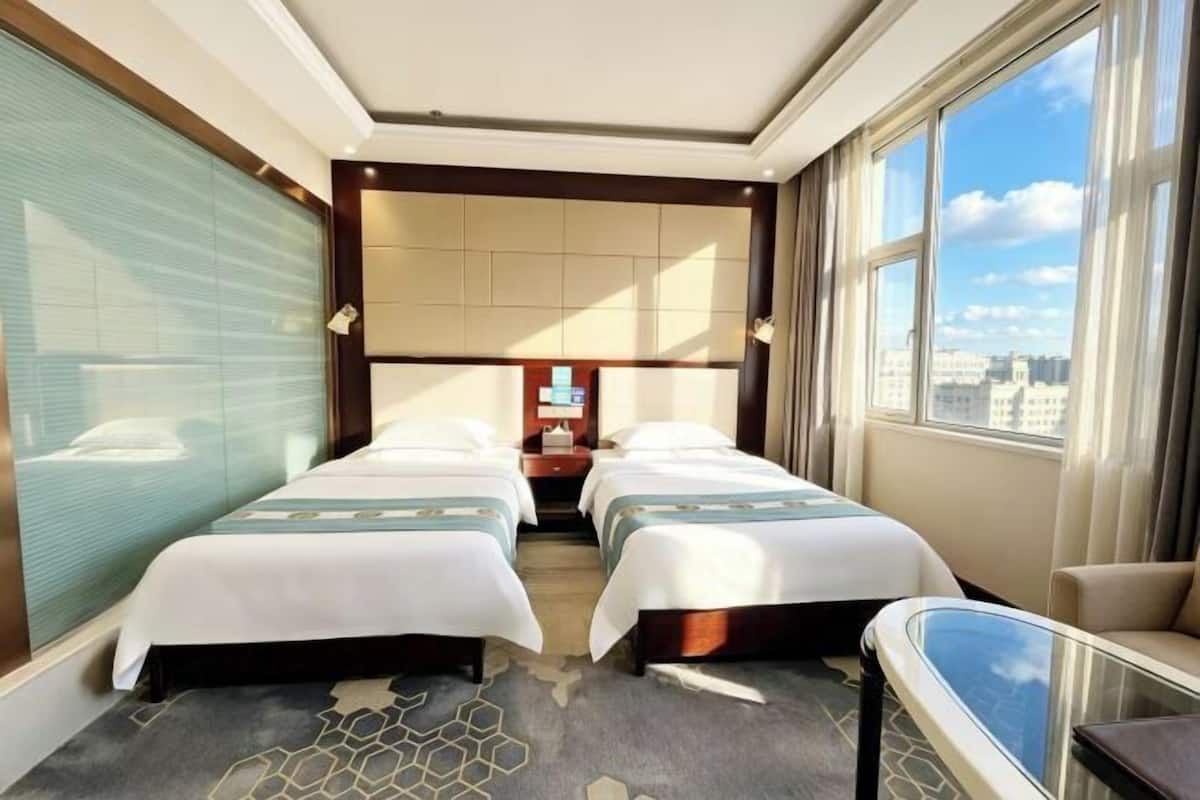A hotel room with two beds, a desk, a chair, and a large window with a city view.