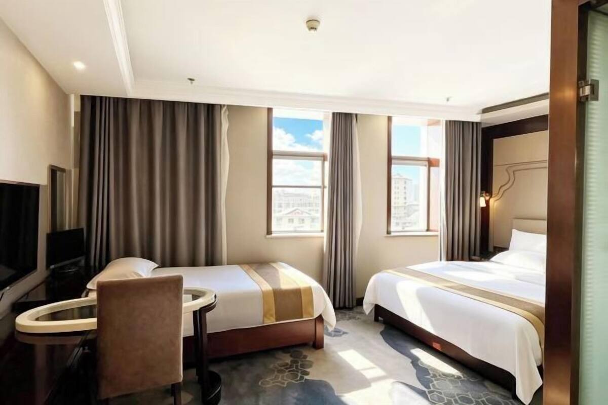 A hotel room with two beds, a desk, a chair, a television, and large windows.