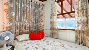 A bedroom with a bed, a red heart-shaped pillow, a fan, and curtains with a floral pattern. - Casa Aconchegante na Lagoa de Ibiraquera (Imbituba)