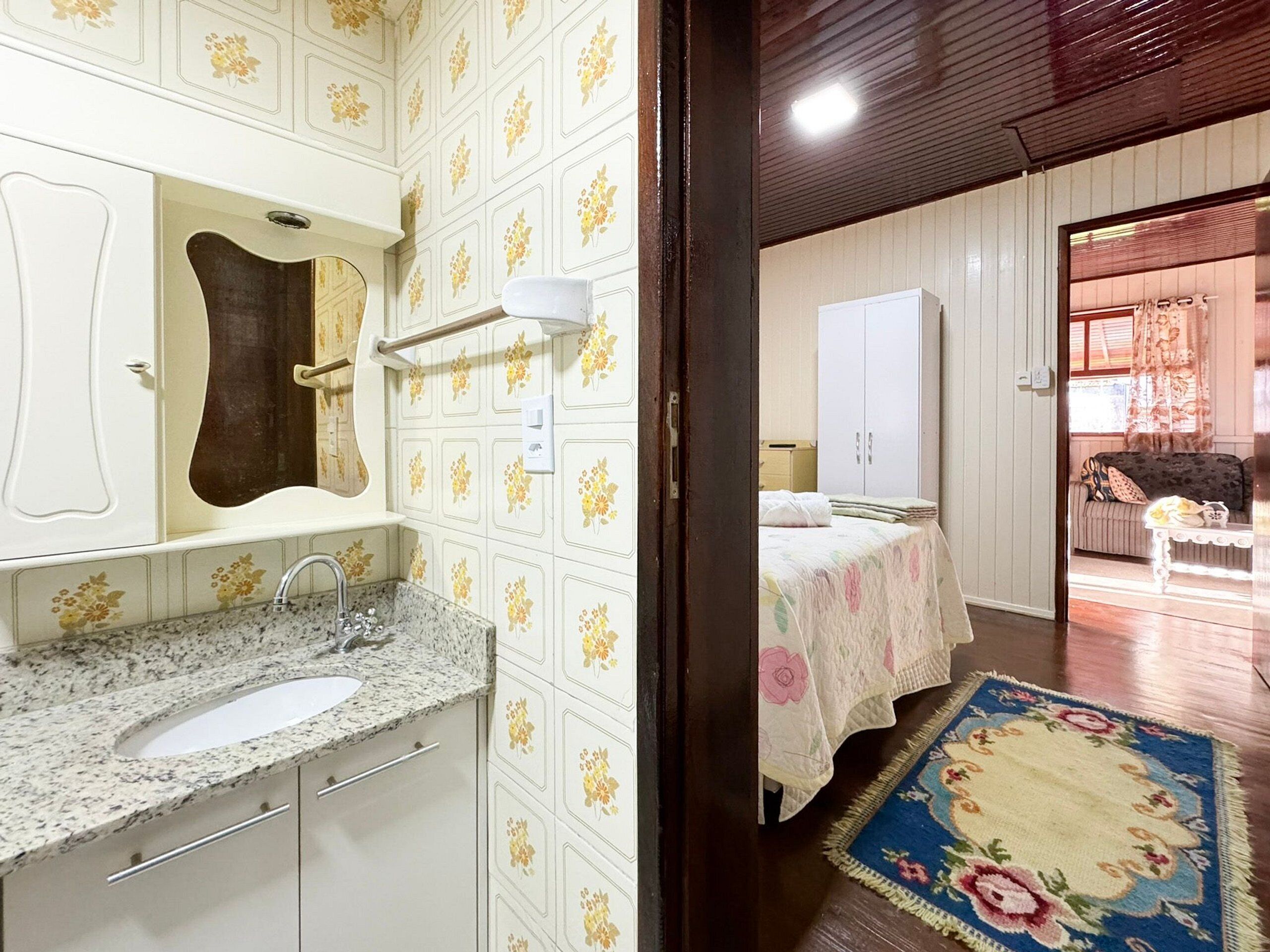 Ein Badezimmer mit Waschbecken, Spiegel und Handtuchhalter. Im angrenzenden Raum befindet sich ein Bett mit floralem Überwurf und ein blaues Blumenmuster-Teppich.