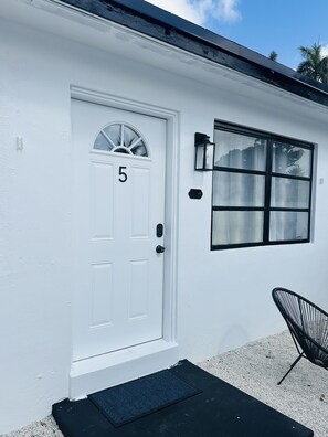 Une maison blanche avec une porte portant le numéro 5, une fenêtre et une chaise noire à l’extérieur.