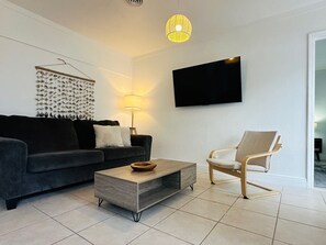 Een moderne woonkamer met een donkergrijze bank, een houten salontafel, een lichtgekleurde fauteuil, een wandgemonteerde flatscreen-tv en een decoratief kunstwerk van vissen aan de muur.