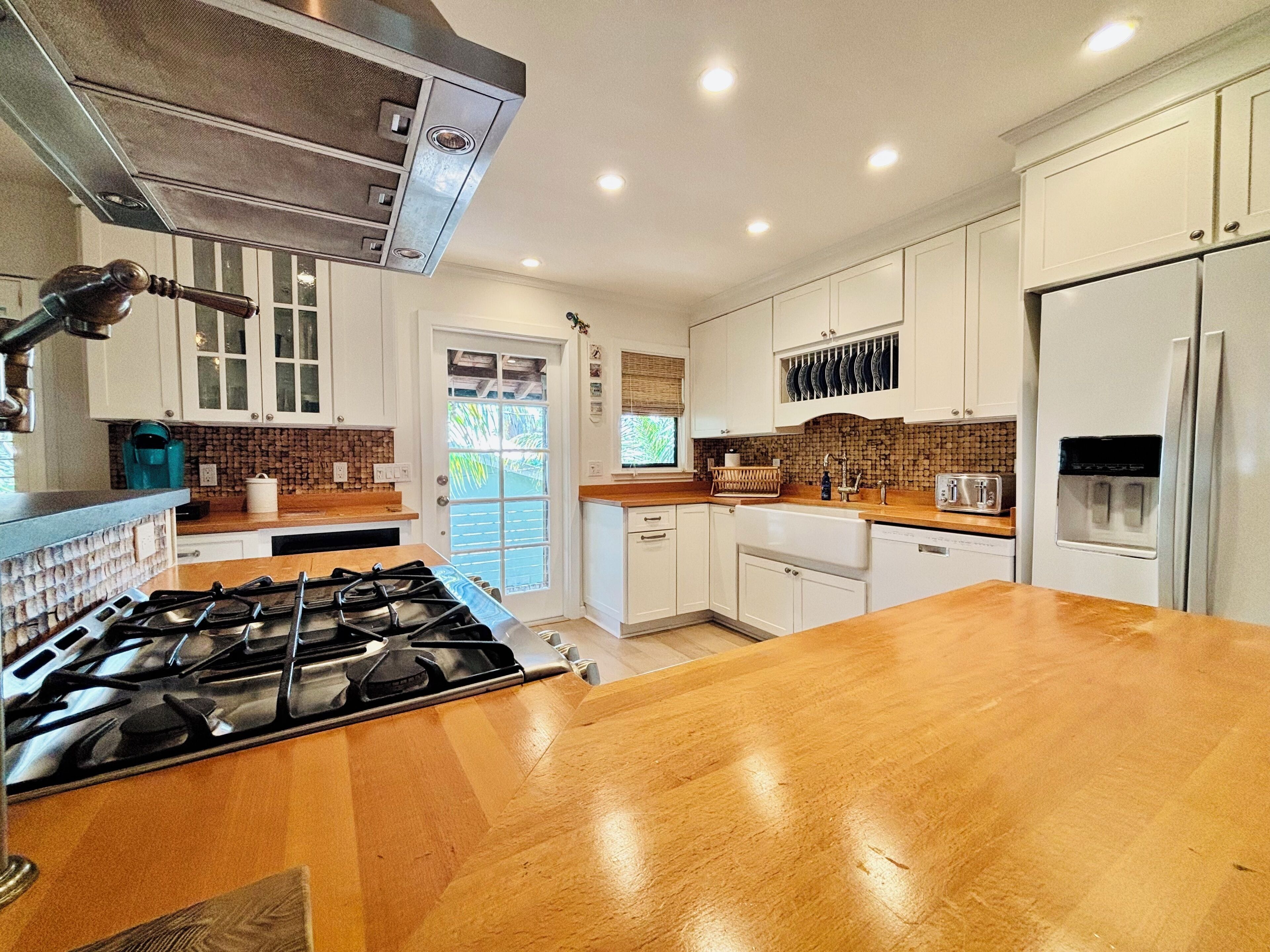 A modern kitchen with white cabinets, a wooden countertop, a gas stove, a refrigerator, and a window with a view of greenery.