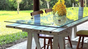 Une table à plateau en verre, entourée de chaises, placée sous une pergola en bois, le tout au milieu d’une végétation luxuriante.