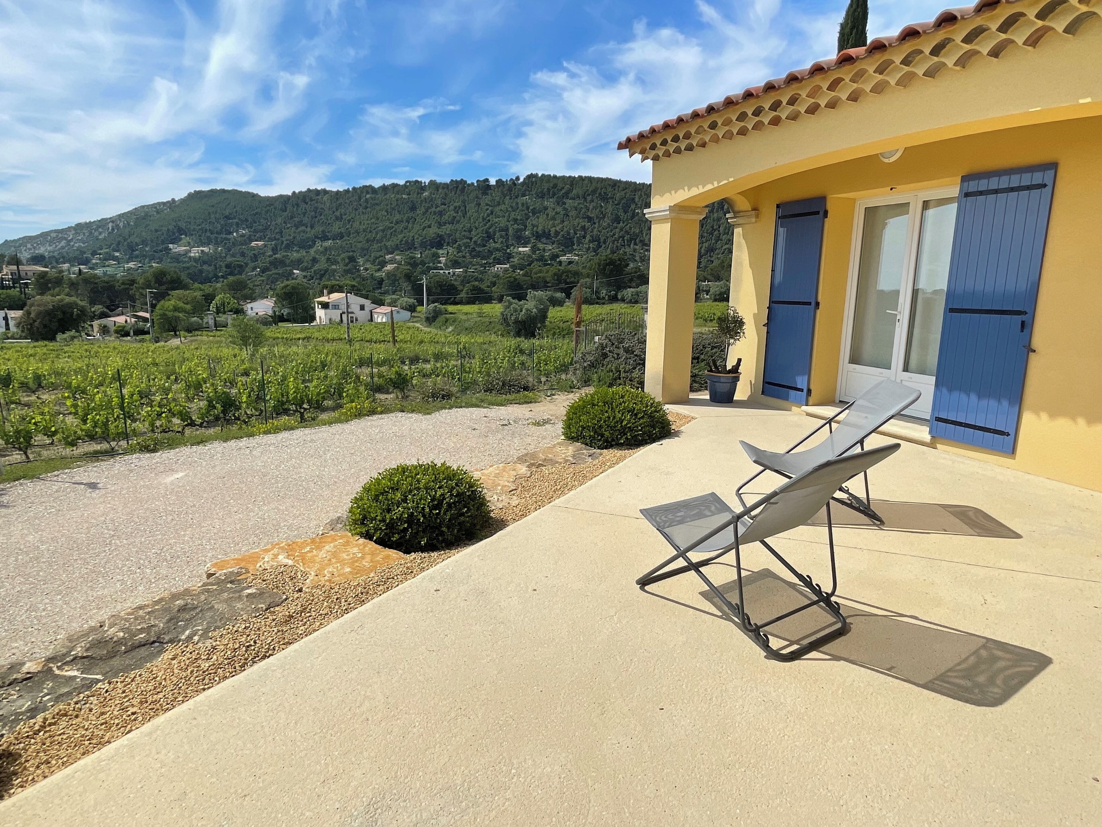 A yellow building with a tiled roof, a covered patio with a table and chairs, and a view of vineyards and hills in the background.