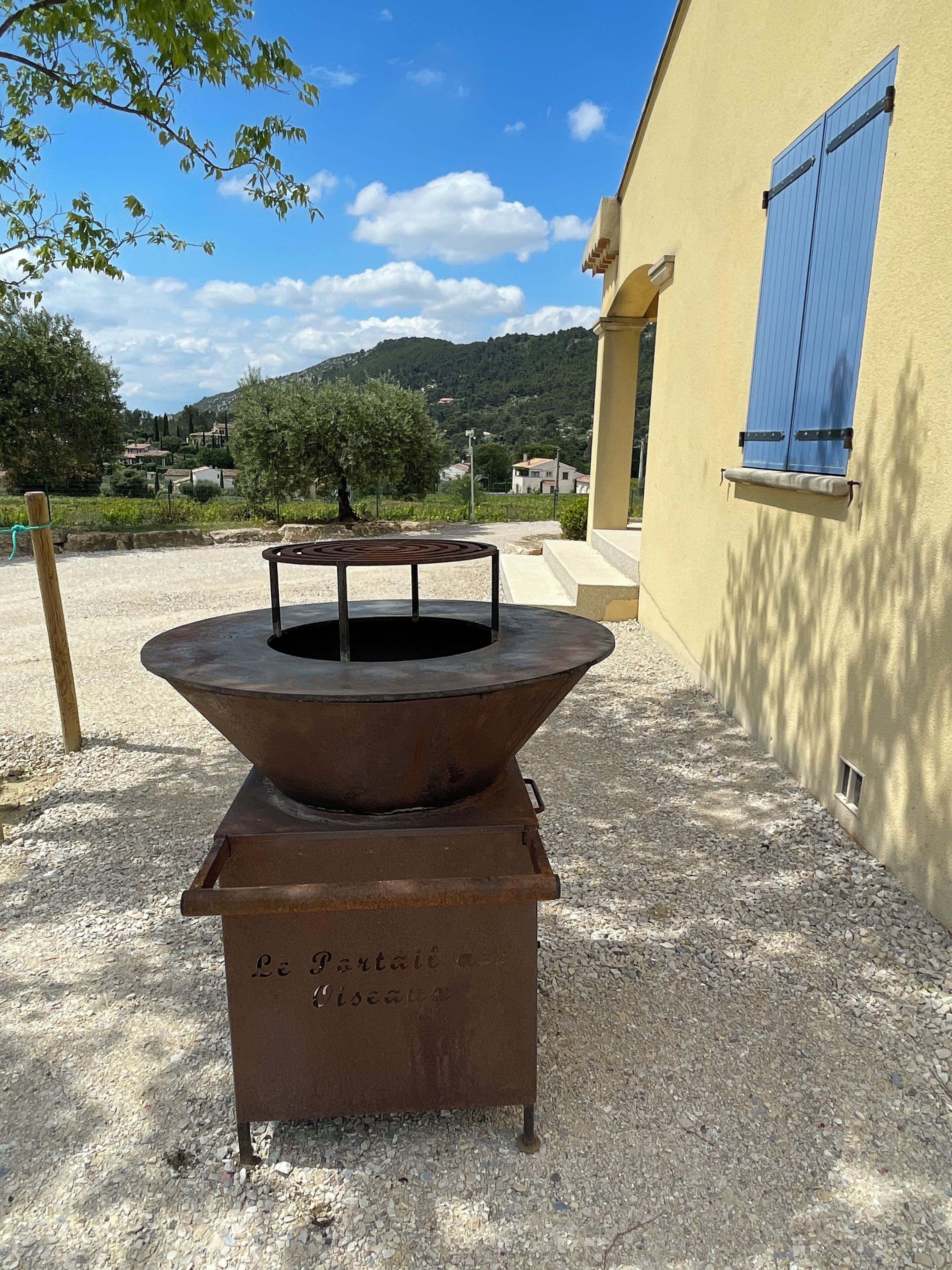 A rustic outdoor fire pit with a metal stand, set on gravel, in front of a yellow building with blue shutters.