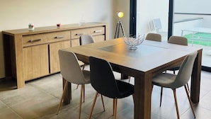 Un espace repas moderne avec une table et des chaises en bois, un luminaire suspendu au-dessus et une vue sur l’extérieur grâce à une grande fenêtre.