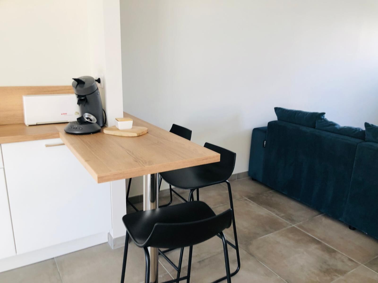 Une petite cuisine équipée d’une machine à café, d’une planche à découper et d’un appareil électroménager blanc. La pièce contient deux chaises noires et un canapé bleu foncé.