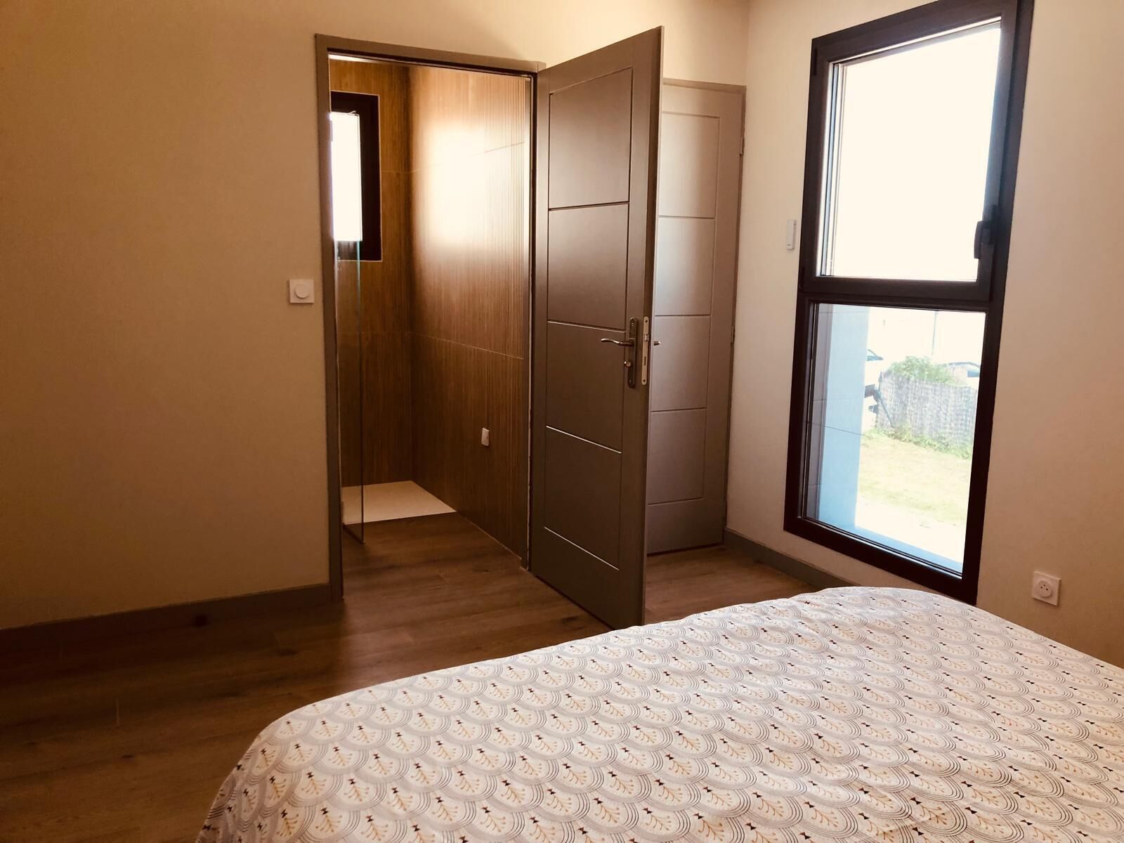 A bedroom with a bed, a wooden wardrobe, a door leading to a bathroom, and a window with a view of the outside.