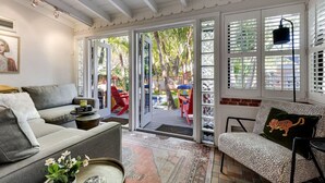 Una sala de estar acogedora con un sofá gris, un sillón estampado y una mesa de centro. Hay un ventanal con persianas, una puerta que da al exterior y una mesita con un jarrón de flores.