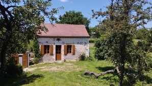A rustic stone house with a red-tiled roof, wooden shutters, and a small front yard.