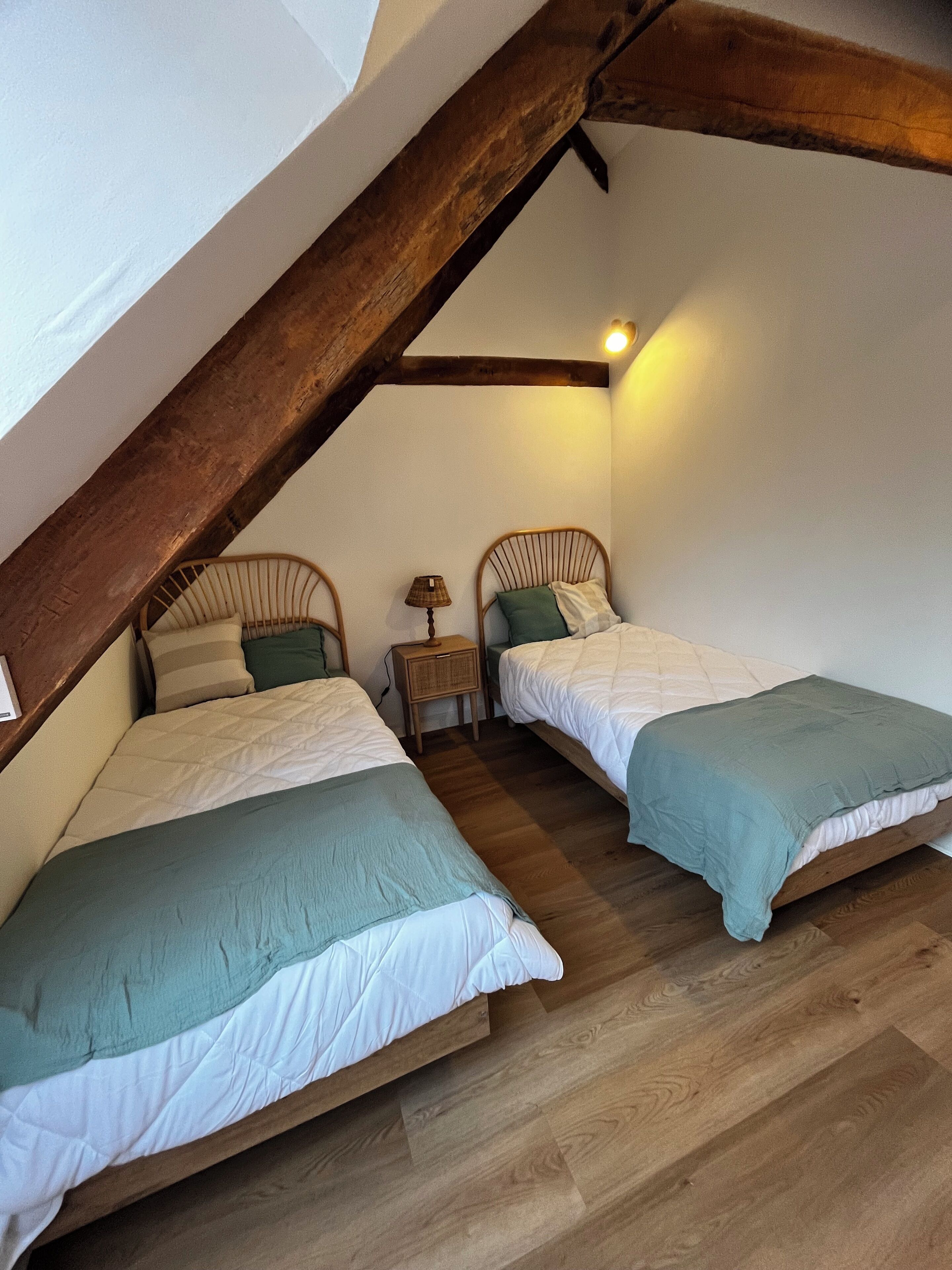 A bedroom with two single beds, wooden beams, a nightstand with a lamp, and a small window.