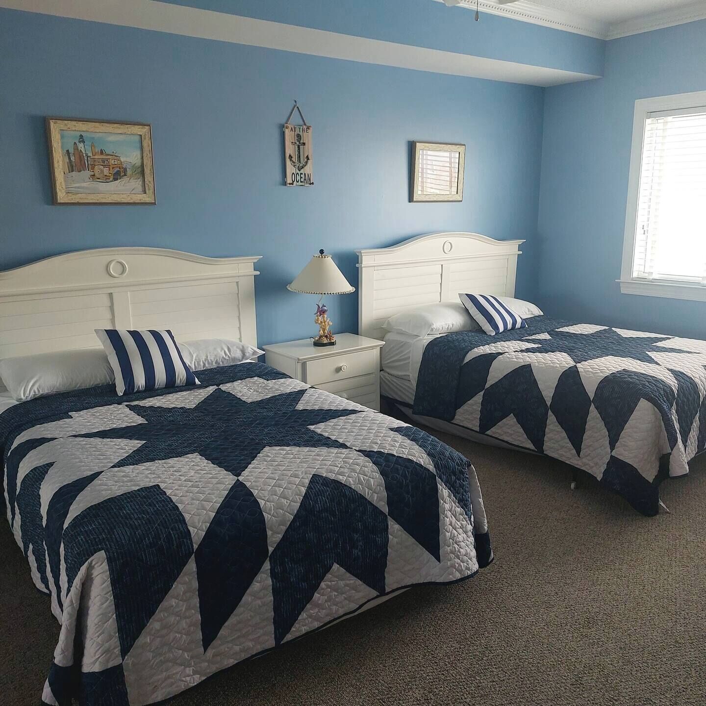 Un dormitorio con dos camas, cada una con colcha azul y blanca, cabeceros blancos y una mesita de noche con lámpara.