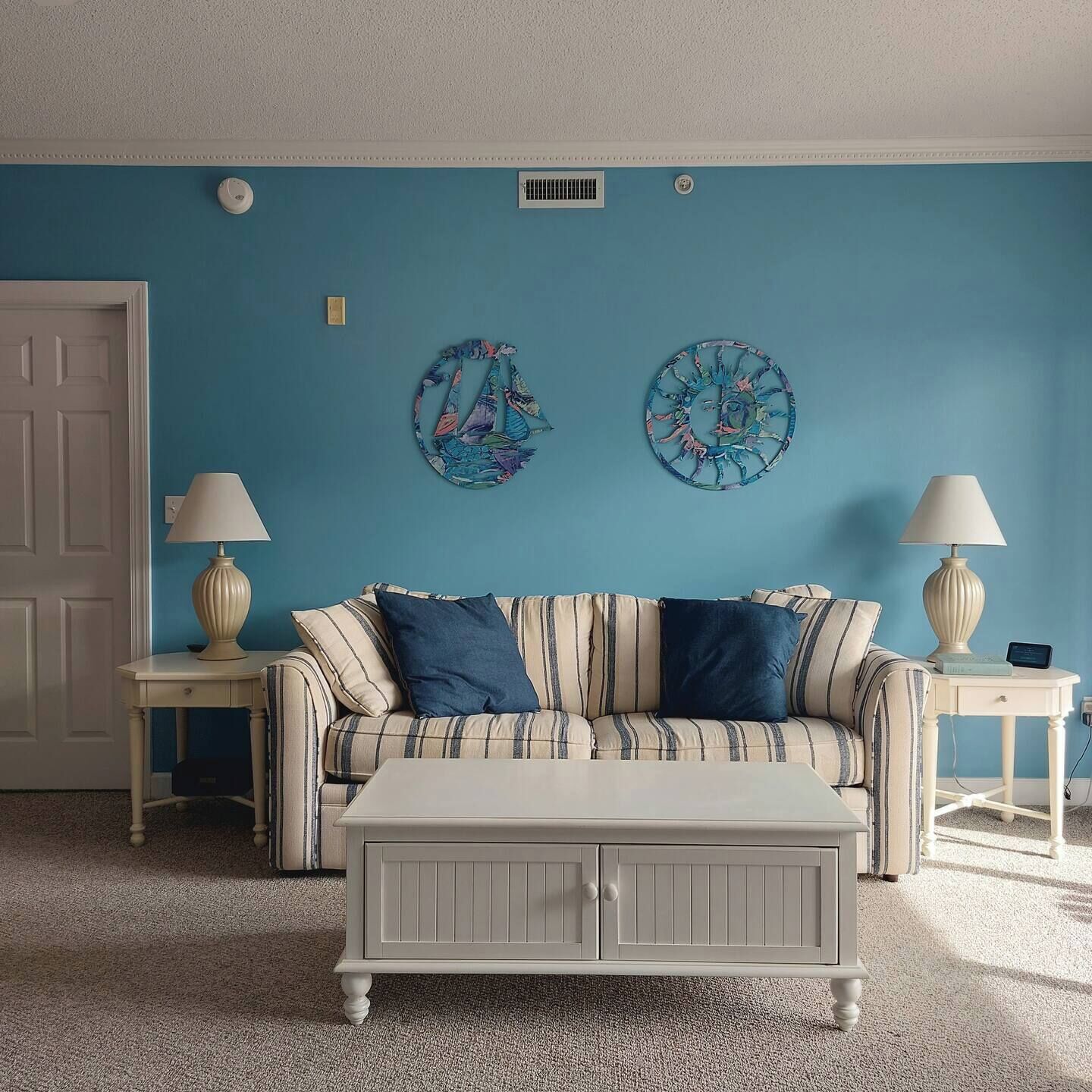Una habitación con una pared azul, un sofá a rayas, dos mesitas de noche con lámparas y una mesa de centro blanca.