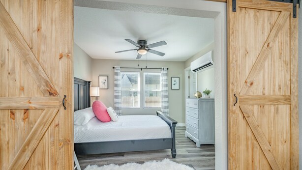 Un dormitorio con cama, ventilador de techo, ventana con persianas, cómoda y una alfombra blanca y mullida.