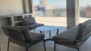 Un espace extérieur aménagé, abrité, avec une table ronde et deux canapés, un barbecue intégré et une vue sur le paysage urbain.