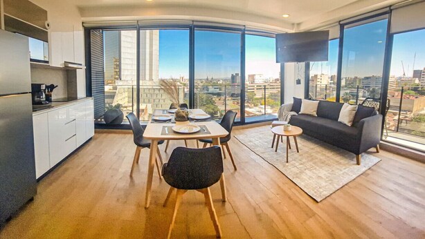A modern living room with a dining area, large windows offering city views, a flat-screen TV, and a kitchen with white cabinets. - New apartment Nous 606 (Guadalajara)